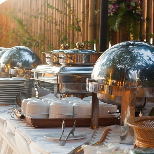 Outdoor buffet table with chafing dishes, stacks of plates, and white bowls.
