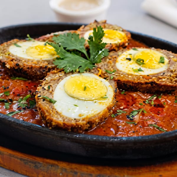Four Scotch eggs, halved, in a sizzling cast iron pan with red sauce and cilantro.