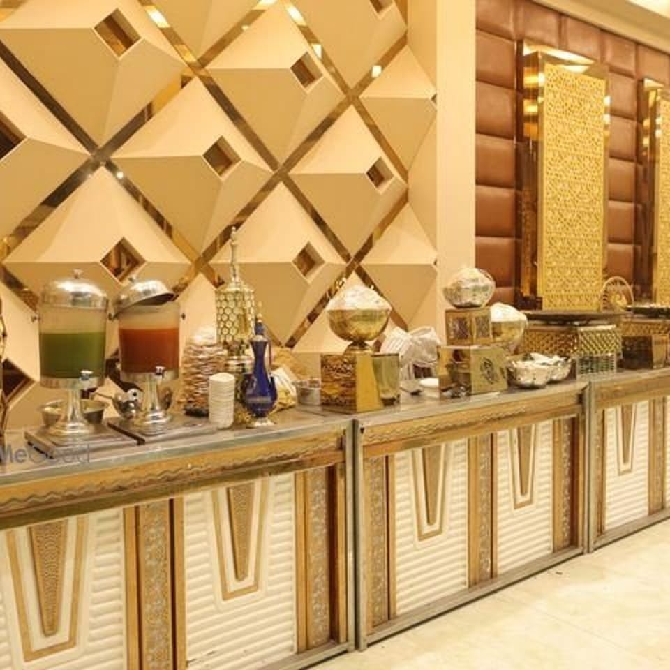 A long, ornate buffet table with various food and drink dispensers, against a geometric gold and cream wall.