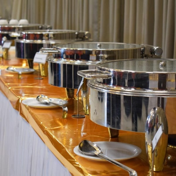 A buffet line with several silver chafing dishes on a gold tablecloth with white skirting.