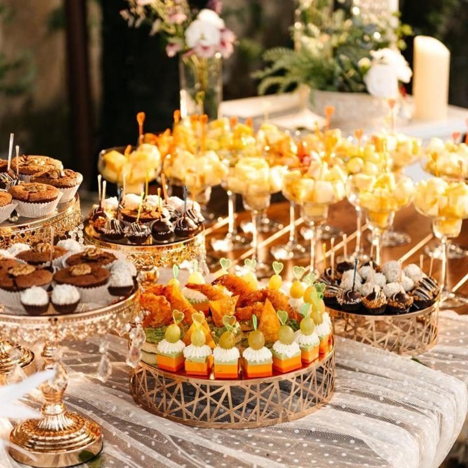 Elegant dessert table laden with cupcakes, chocolate treats, fruit cups, and savory appetizers on golden stands.