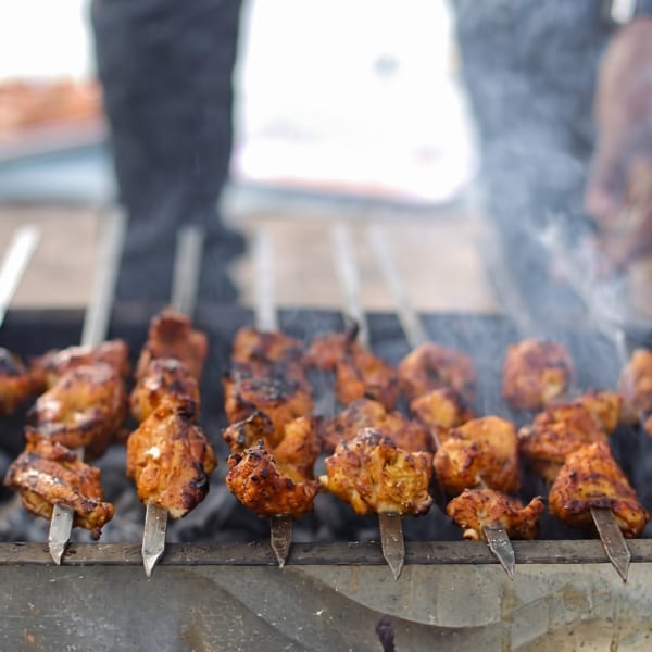 Chicken tikka skewers grilling over hot coals with smoke rising, a person's hands visible in the background.