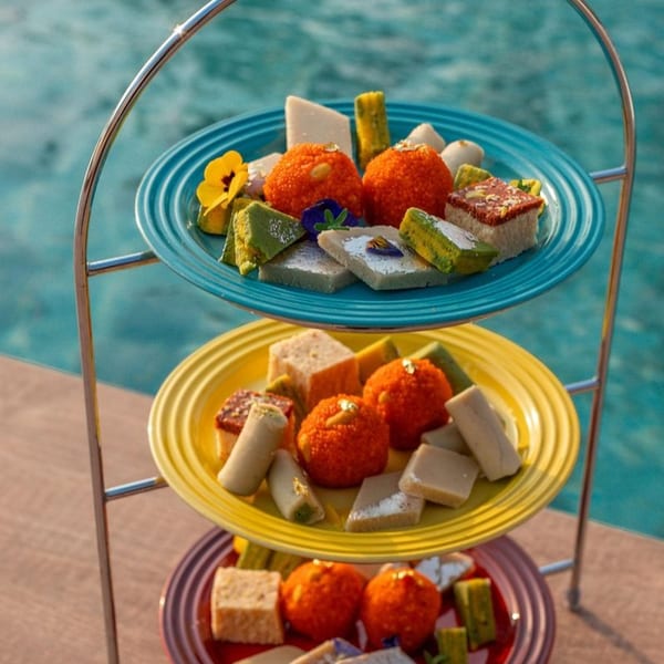 A three-tiered stand holds colorful Indian sweets, including ladoo and barfi, by a swimming pool.
